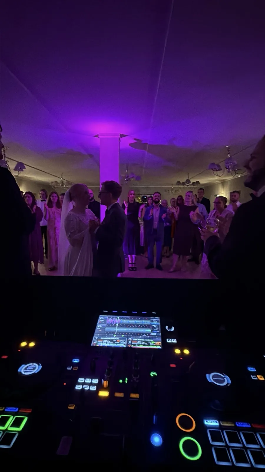 Brudeparret danser brudevals midt på gulvet, set bag DJ-pulten. Bruden i hvid blondekjole og slør, gommen i mørkt jakkesæt. Rundt om dem står gæsterne i en kreds og ser på, under et hvidt loft med en søjle i lilla lys og lysekroner langs væggene. Forrest lyser pultens skærm og knapper.